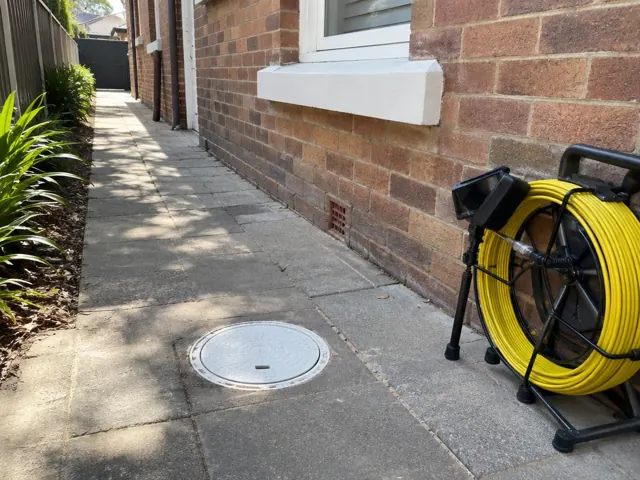 Capped drain inspection opening on a paved side path beside a brick house, with a coiled camera reel in Ashfield