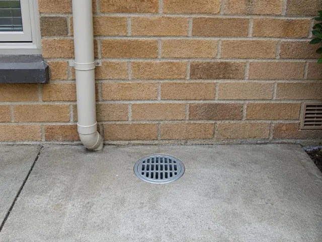 Outdoor drain grate set in dry concrete beside a brick wall and downpipe in Croydon