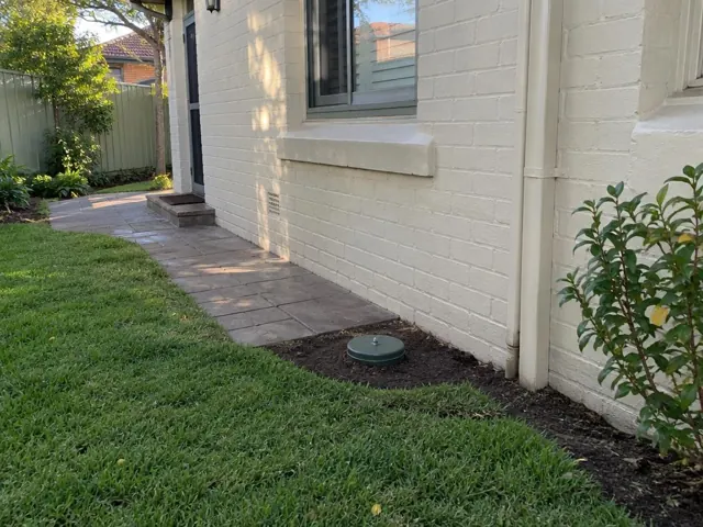Capped sewer opening in a narrow garden bed beside a cream brick house in Five Dock